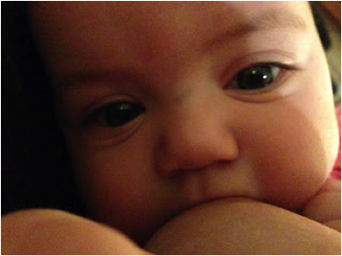 Close up face shot of a white infant with large green eyes latched onto her mother’s breast