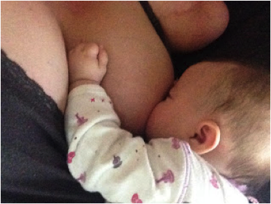 White woman amputee is shown breastfeeding white infant with dark brown fuzzy hair in pink and purple pajamas. Infant latched onto her mother’s left breast and her left hand rests between the mother’s breasts