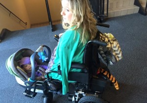 White woman with no legs and one arm in power wheelchair with baby carseat/carrier wedged between wheelchair footrests.