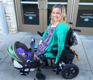 White woman with no legs and one arm smiling at camera in power wheelchair with baby carseat/carrier snugly wedged between wheelchair footrests. Baby is facing the woman.