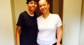 pic of daughter and mother standing together in her dorm room. She's wearing black t-shirt and shorts with a hat turned backwards. mom is wearing white t-shirt and brown sweatpants while holding onto cane.