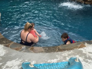 White woman triple amputee sitting in pool holding baby with young boy next to them wearing life vest