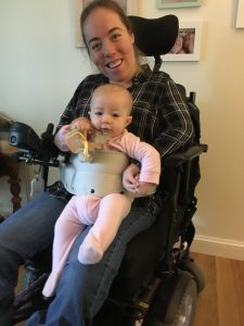 Photo of woman in wheelchair holding baby on her lap. Strap is around her and baby.