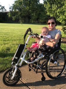 Mom sits in wheelchair with an attachment connected to the front with a motor. Baby is secured on mom's lap with a wide strap. Baby and mom are smiling outside.