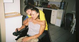 Latina woman in a power wheelchair with son standing behind with his arm wrapped around her.