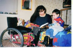 Woman sitting on bed reading book to young boy. Manual wheelchair is next to bed.