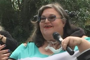 Image of Carrie Ann, a white woman in a wheelchair speaking into microphone 