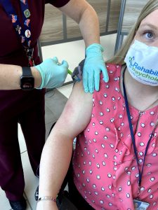 White arms and gloved hands of nurse in scrubs injecting author's arm with vaccine. Author wearing pink polka dotted top and mast.